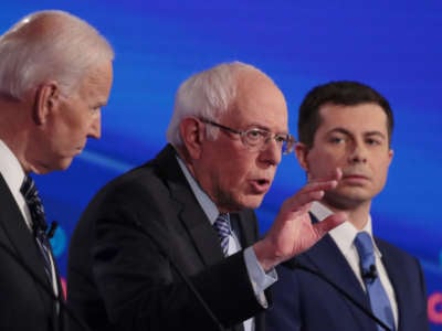 Bernie Sanders speaks during a debate while Joe Biden and Mayor Pete stand on either side of him at their respective podiums