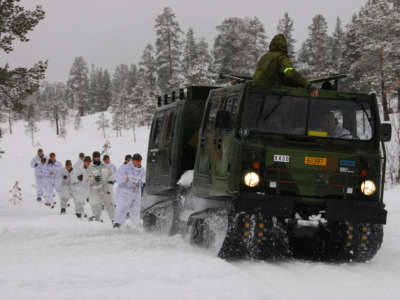 Soldiers dressed in white jog behind a military vehicle driving in the snow