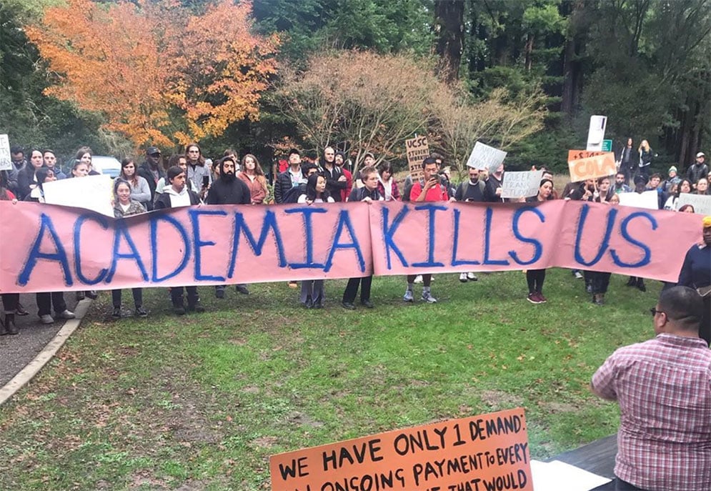 A large group of people hold a sign reading "academia is killing us" outside humanities buildings at University of California Santa Cruz, December 12, 2019.