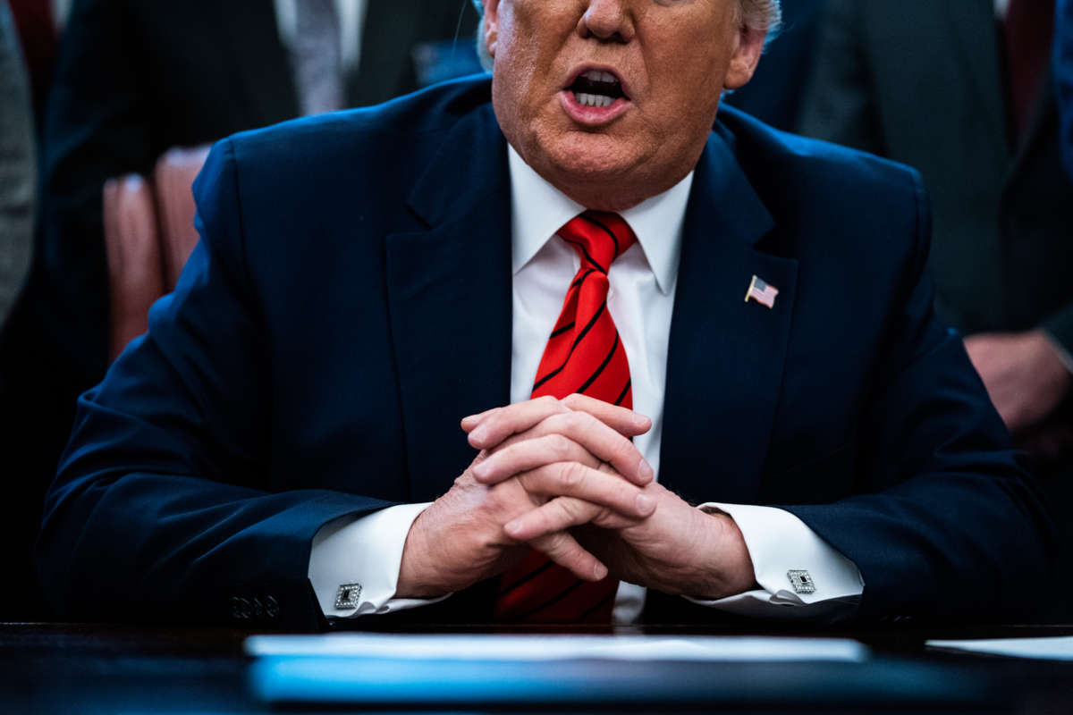 Donald trump, whose folded hands are several shades lighter than his face, speaks while seated at a desk