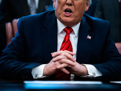 Donald trump, whose folded hands are several shades lighter than his face, speaks while seated at a desk