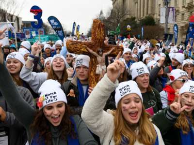 white women in white hats march down the street carrying a cross