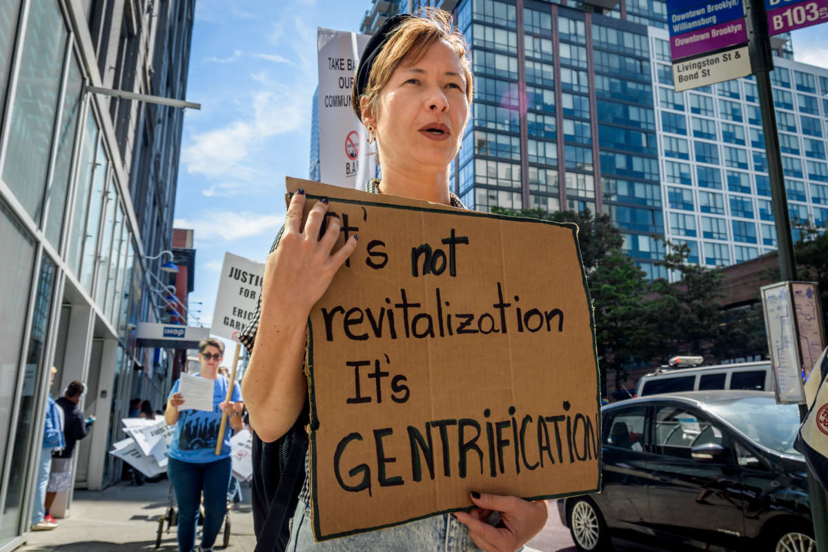 A woman holds a sign reading "It's not revitalization it's GENTRIFICATION" written on cardboard