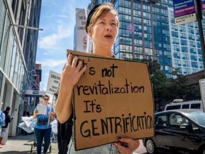 A woman holds a sign reading "It's not revitalization it's GENTRIFICATION" written on cardboard