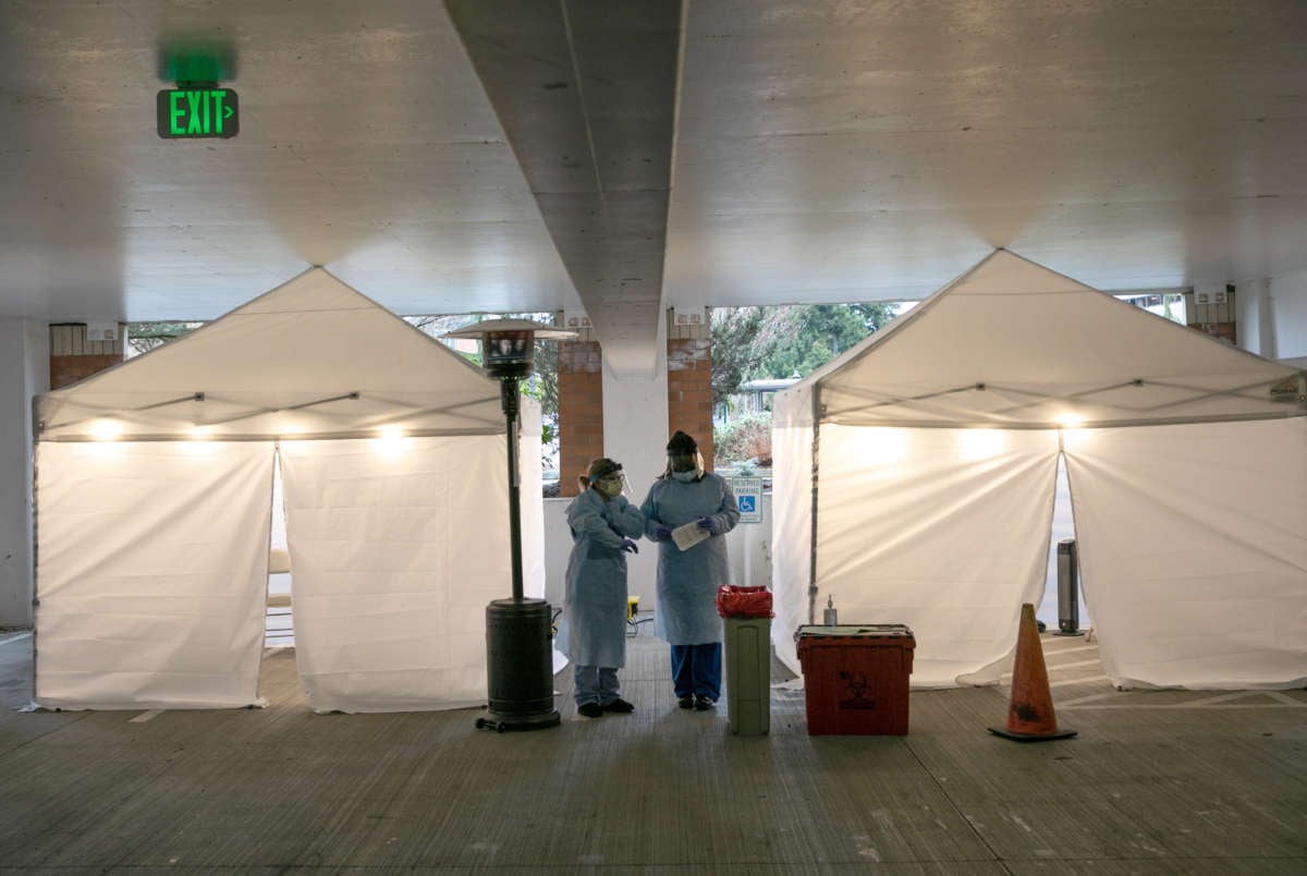 Two medical professionals in scrubs and masks stand between two tents at a drive-through human coronavirus testing station