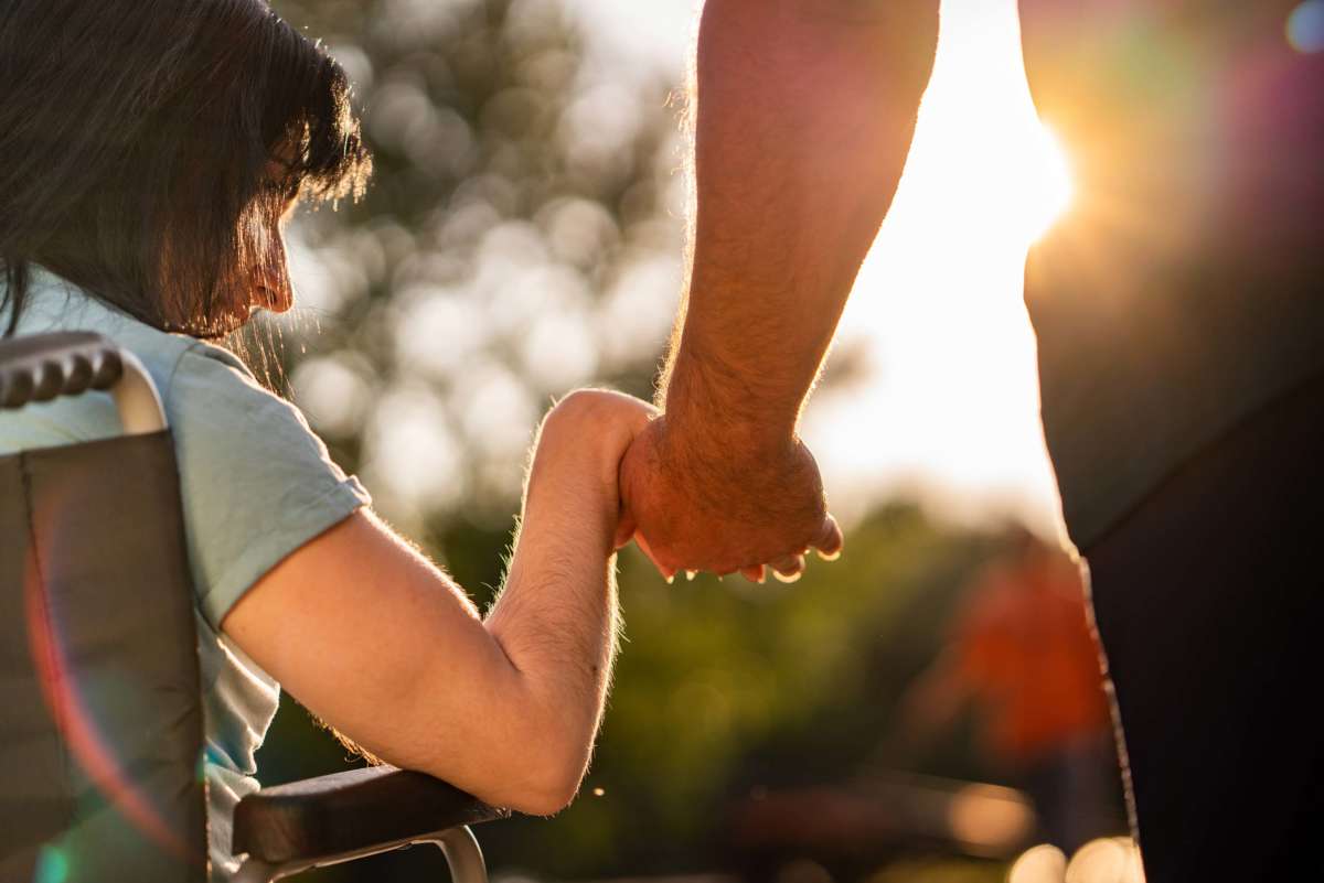 A person in a wheelchair holds hands with another while sun shines