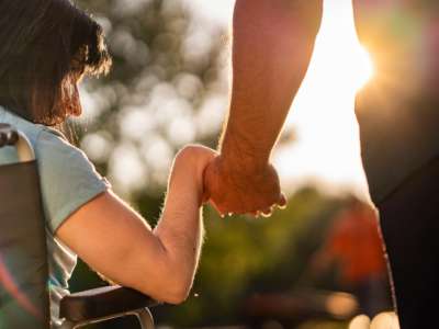 A person in a wheelchair holds hands with another while sun shines