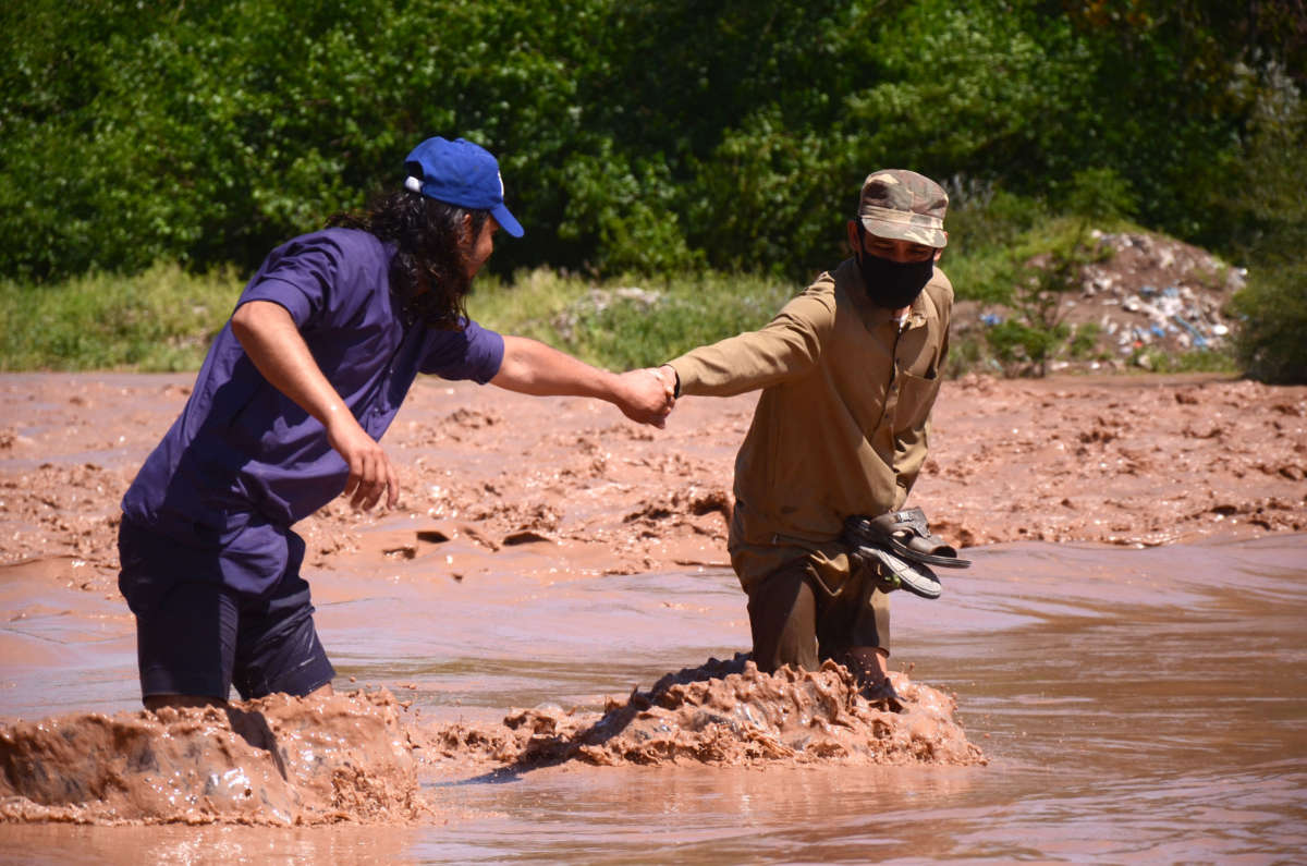 A man in a mask leads another through turbulent flood waters