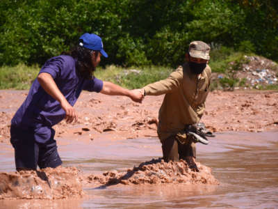 A man in a mask leads another through turbulent flood waters