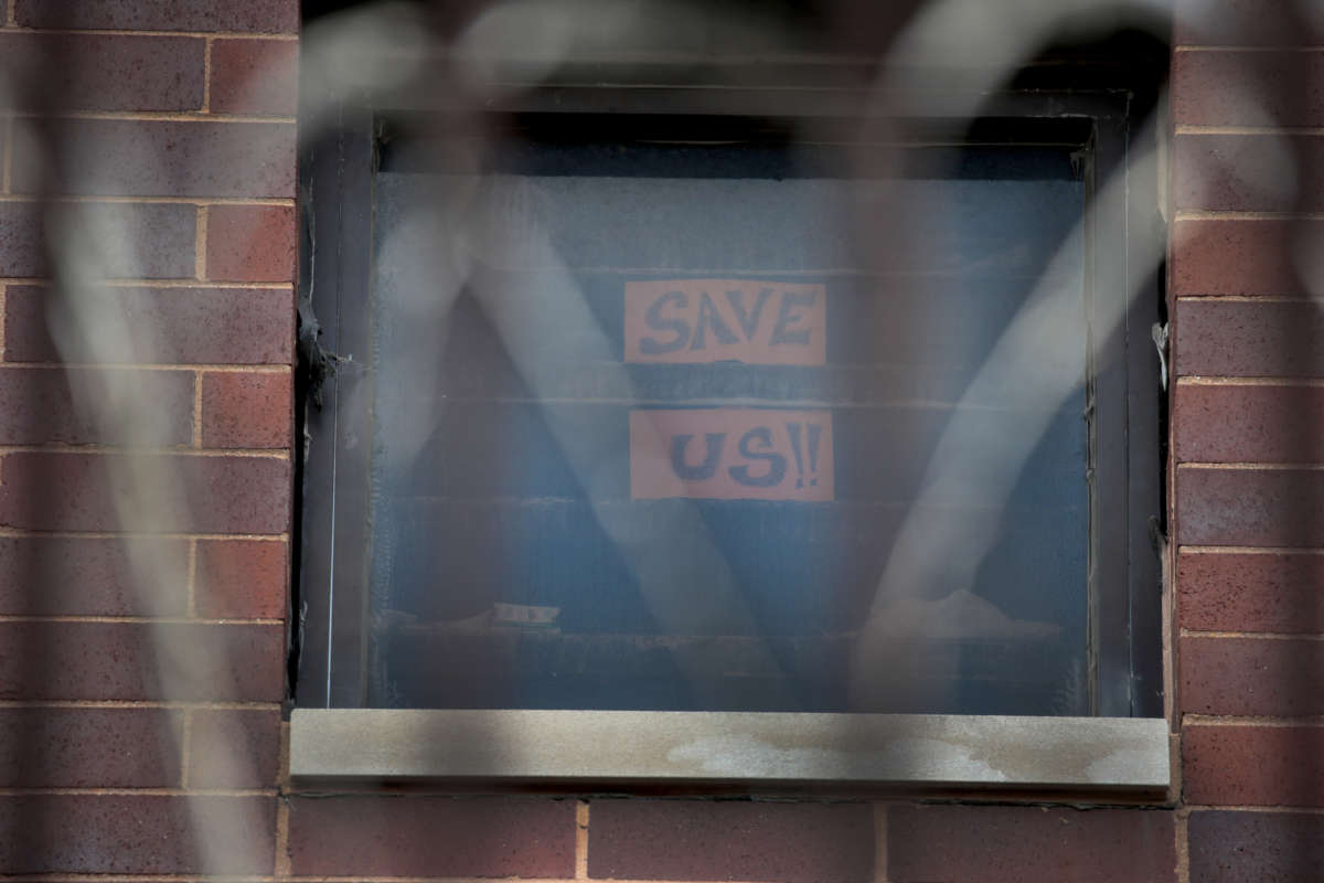 A sign reading "SAVE US" is displayed in a prison window festooned with razor wire