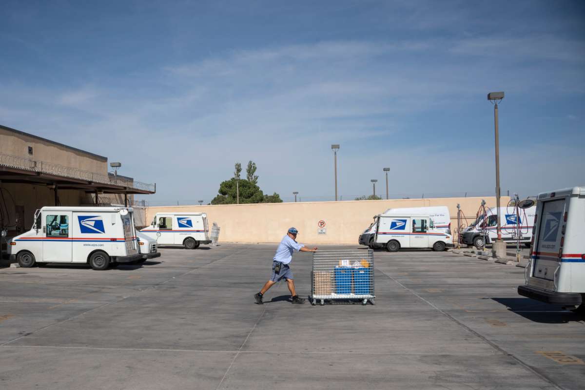 A postal worker pushes a cart towards a mail truck in a parking lot sparsely populated with mail trucks