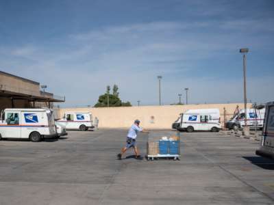 A postal worker pushes a cart towards a mail truck in a parking lot sparsely populated with mail trucks