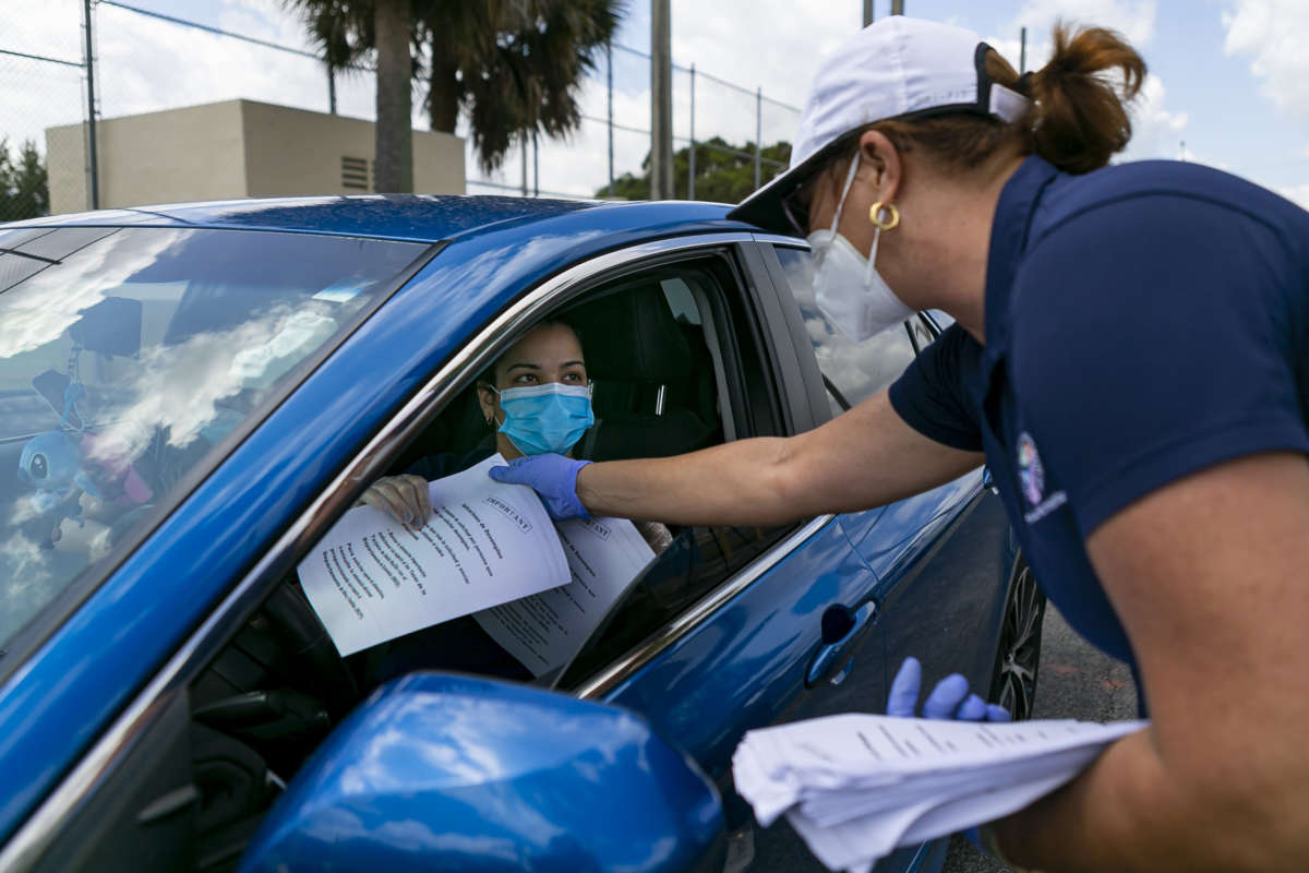 A woman in a protective face mask hands a similarly masked woman in a car some paper