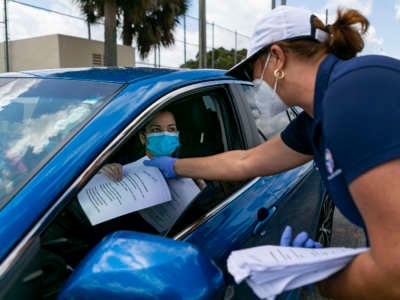 A woman in a protective face mask hands a similarly masked woman in a car some paper