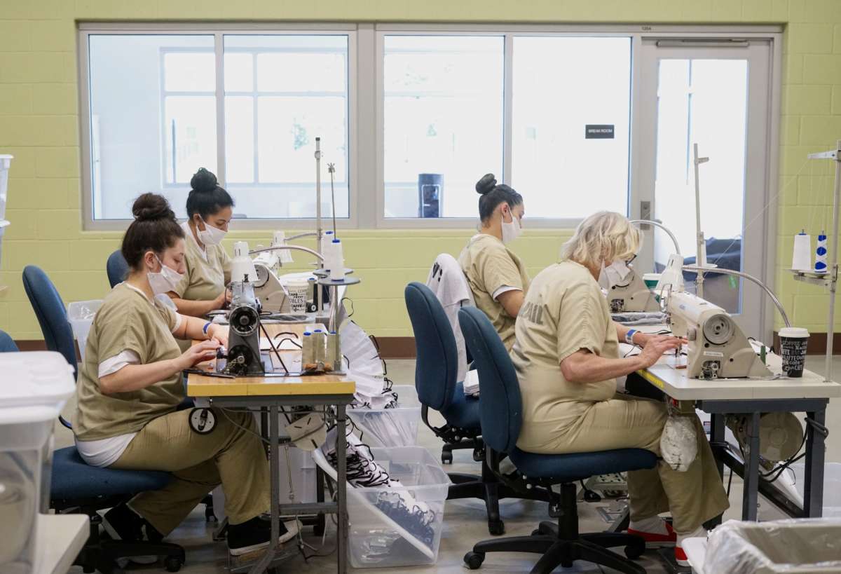 Four women in two rows construct face masks at workstations reminiscent of a sweatshop