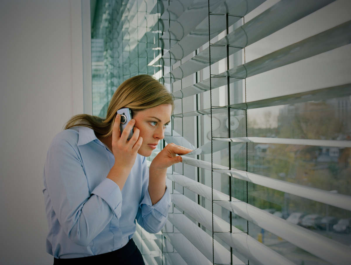 A white woman looks through her blinds while on the phone
