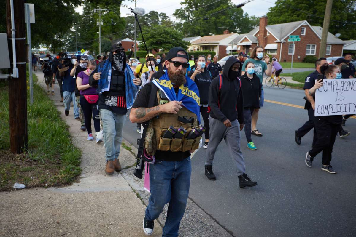 A bearded man carrying a gun walks menacingly beside peaceful protesters