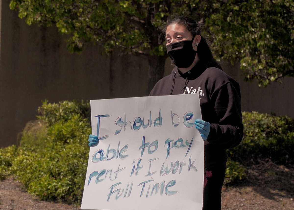 a woman displays a sign reading "I should be able to pay rent if I work full time" while wearing a face mask