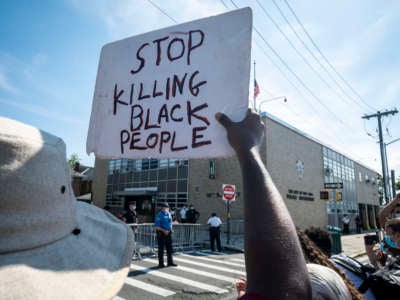 A protester holds a sign that says, "Stop Killing Black People" as the crowd stands in front of the 111th New York Police Department Precinct with police officers looking on during the Black Lives Matter protest in Bayside, Queens, NYC, on August 1, 2020.