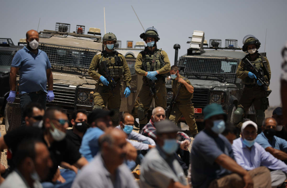 Palestinians kneel on the ground in prayer while being closely surveiled by armed soldiers and military vehicles