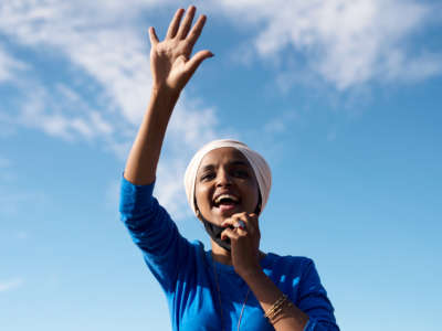 Ilhan Omar waves her hand against a blue sky