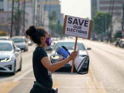 A protester crosses a street holding a sign reading "SAVE OUR ELECTIONS"