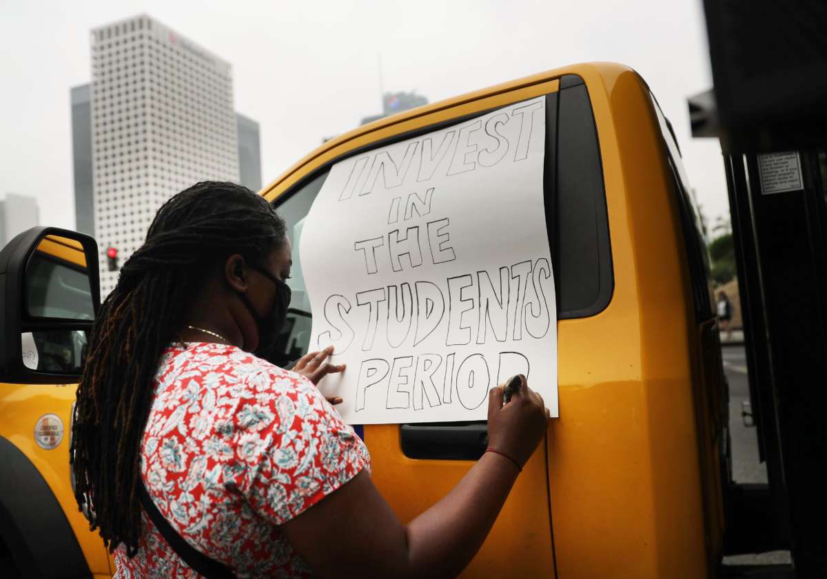 A woman writes a protest sign reading "INVEST IN THE STUDENTS PERIOD"