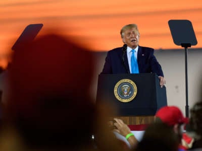 Donald Trump, sweating profusely and matching the orange sunset behind him, speaks at a podium