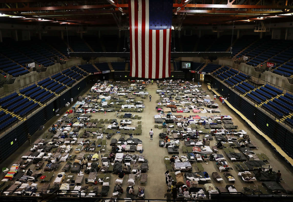An arena bearing a huge us flag houses people sleeping on cots