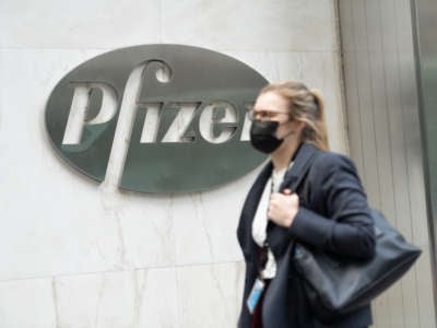 A woman wearing a face mask walks past a Pfizer logo at their Headquarters. Pfizer's coronavirus vaccine could be given to Americans before end of the year, the CEO says.
