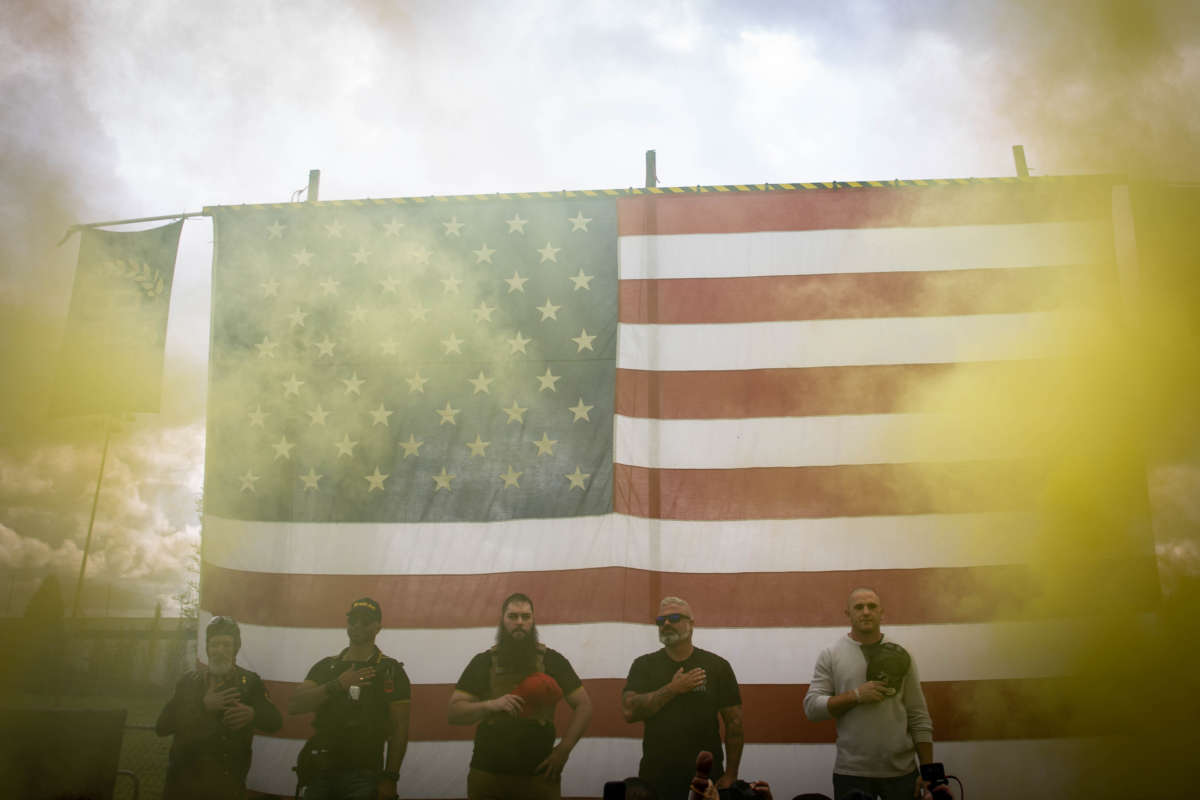 White men hold their maga hats over their hearts as they stand in front of a us flag, yellow smoke billowing in the foreground