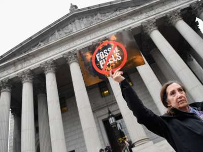 a woman holds a sign reading "[no] FOSSIL FUELS" in front of the new york supreme court building