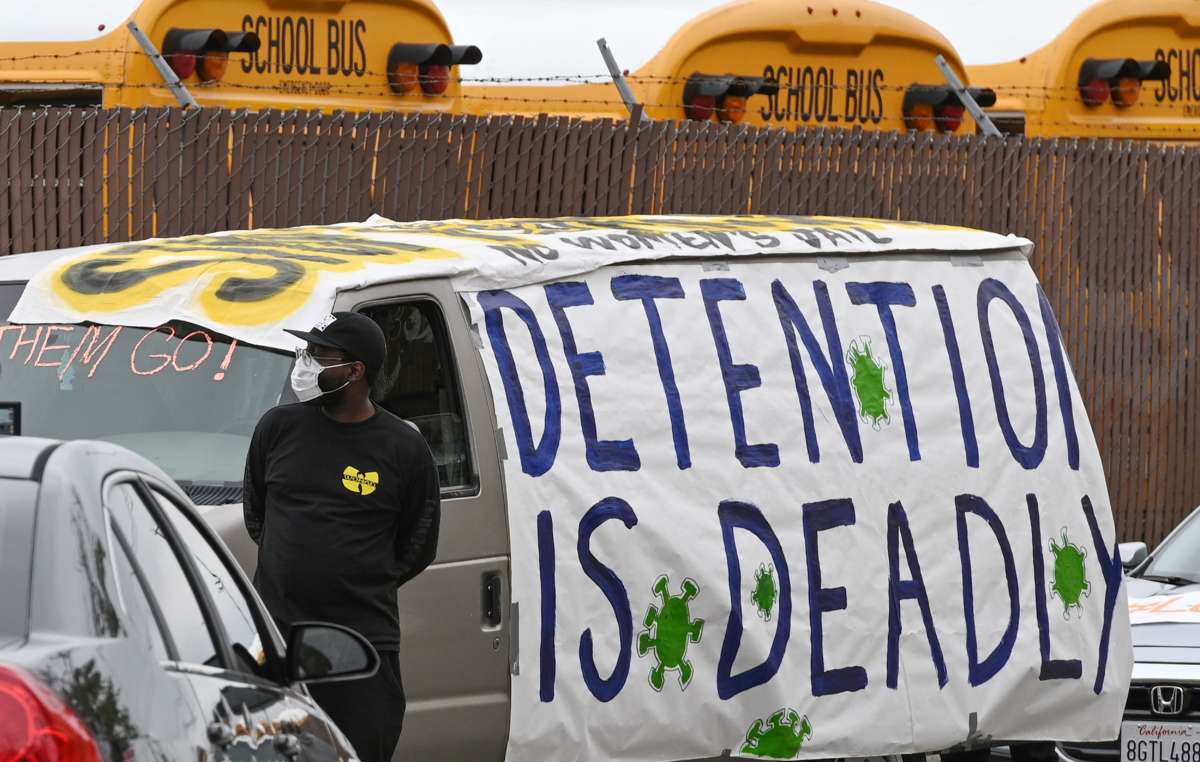 A masked person stands in front of a van covered in a sign reading "DETENTION IS DEADLY"