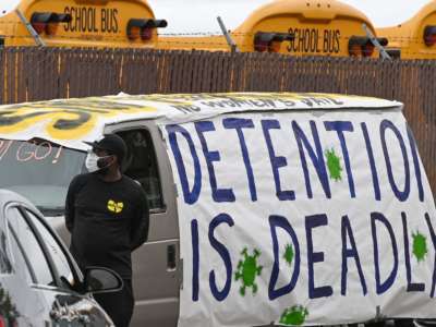 A masked person stands in front of a van covered in a sign reading "DETENTION IS DEADLY"
