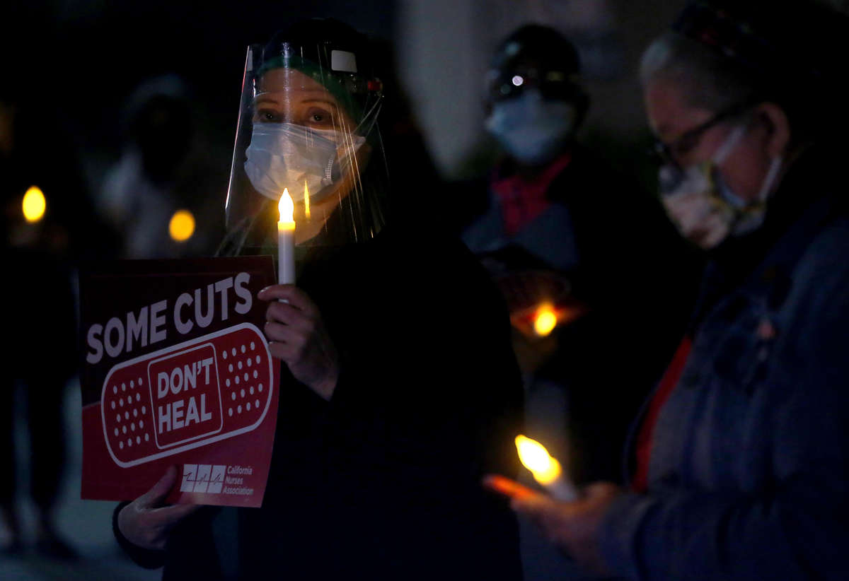 Nurses hold a candle light vigil, one holding a sign reading "SOME CUTS DON'T HEAL"