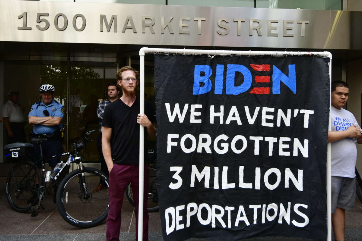 Two activists display a banner reading "BIDEN: WE HAVEN'T FORGOTTEN 3 MILLION DEPORTATIONS"