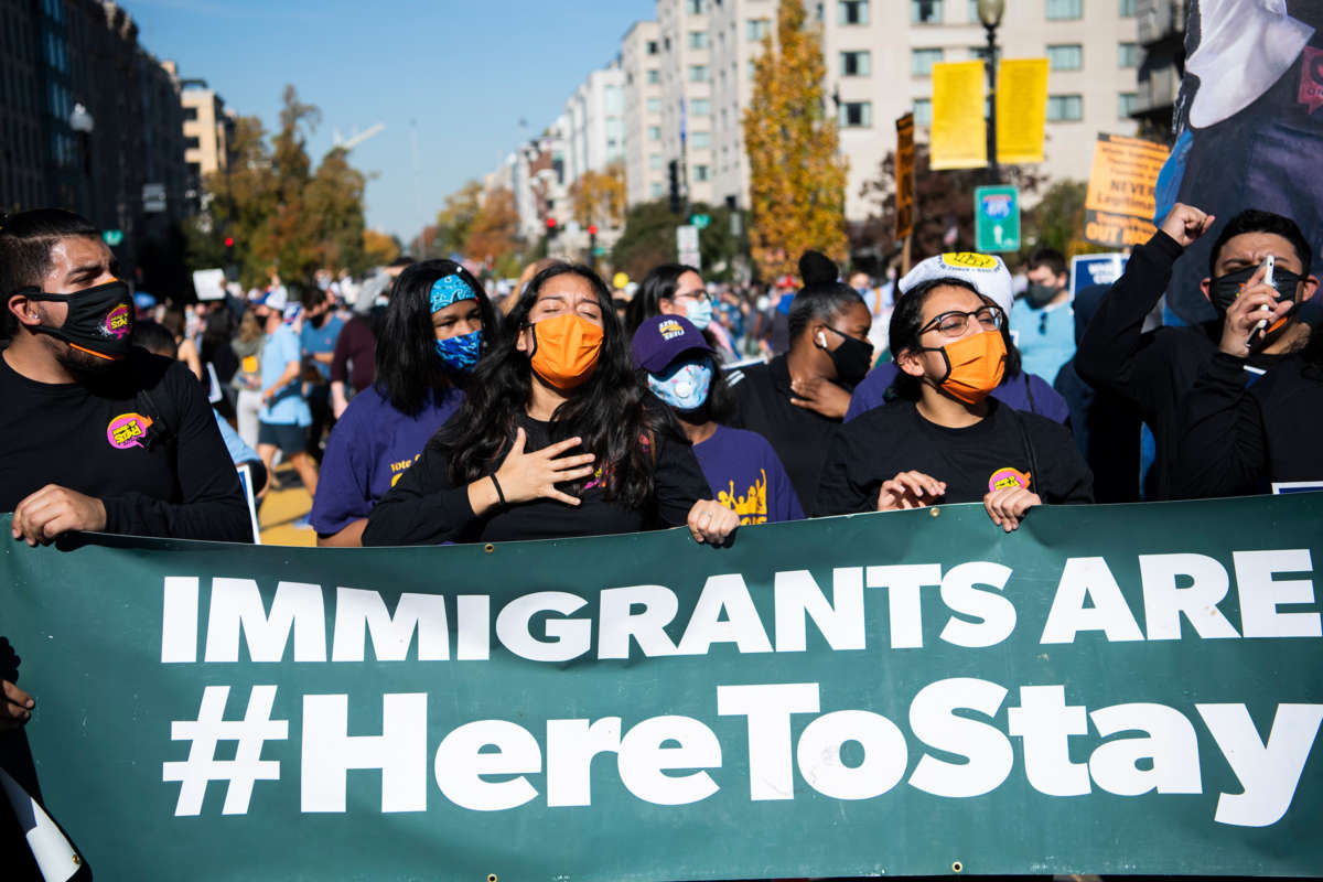 activists march behind a banner reading "IMMIGRANTS ARE HERE TO STAY"