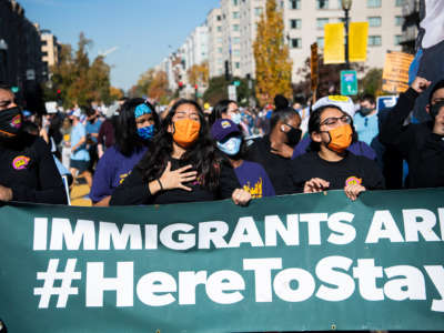 activists march behind a banner reading "IMMIGRANTS ARE HERE TO STAY"