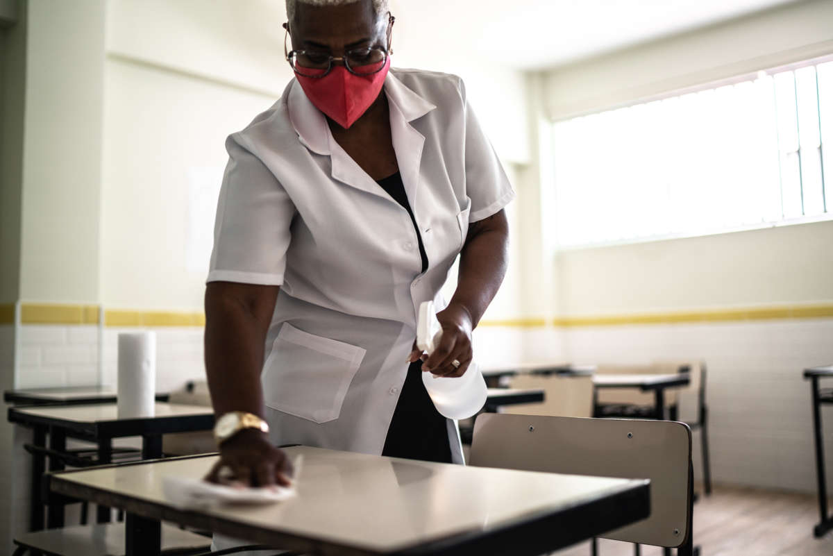 A masked woman cleans a school desk with a spray bottle and cloth