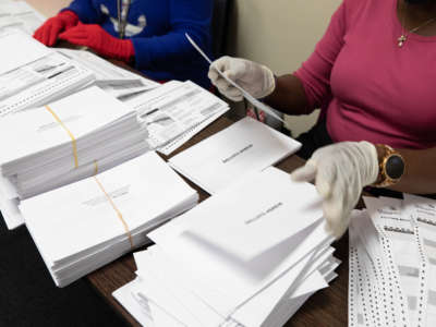 A ballot counter handles a piles upon piles of paper ballots