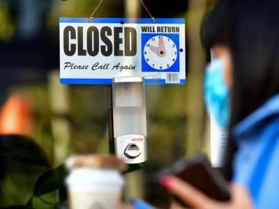 A pedestrian wearing her facemask and holding a cup of coffee walks past a closed sign hanging on the door of a small business in Los Angeles, California, on November 30, 2020.