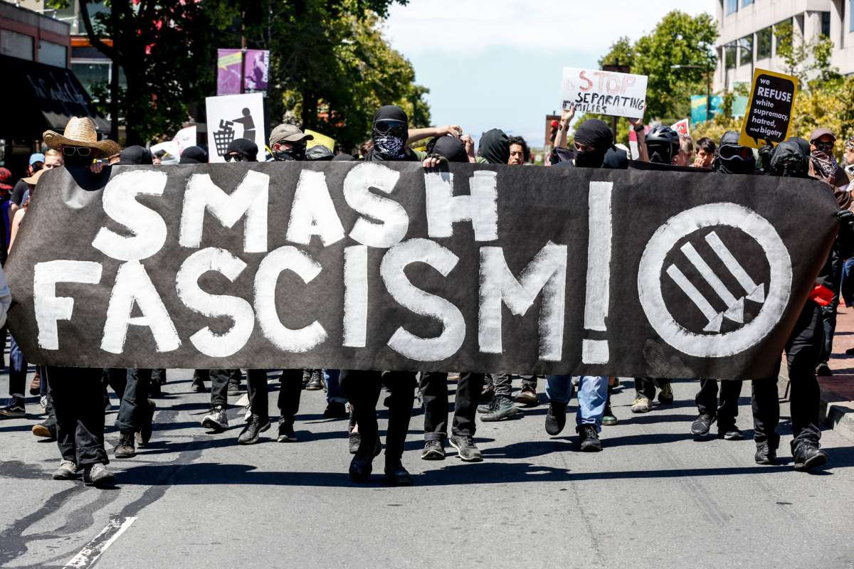 Antifa members march down a street behind a banner reading "SMASH FASCISM"