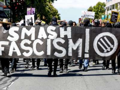 Antifa members march down a street behind a banner reading "SMASH FASCISM"