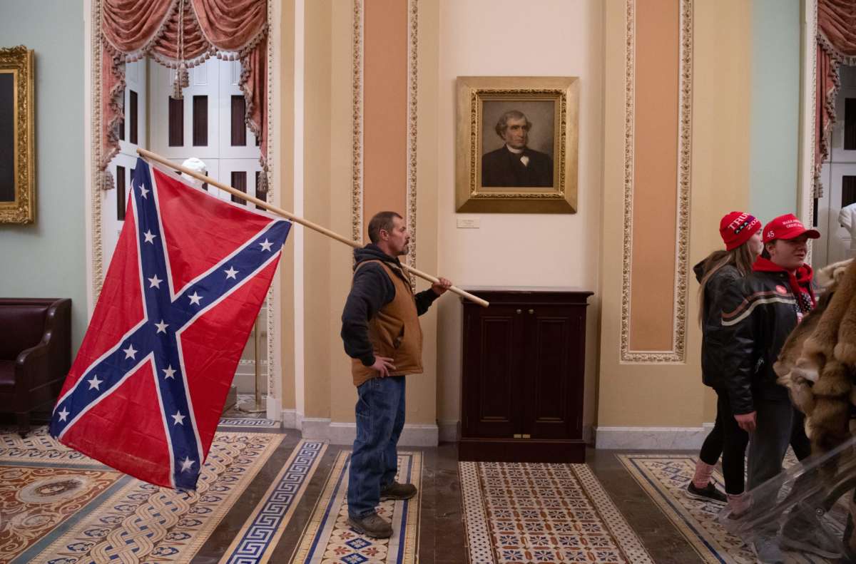 A white man, inexplicably still alive after storming the U.S. capitol building, holds the flag of a defunct seditionist government