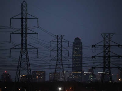 A darkened texas skyline stands behind electrical towers in the foreground