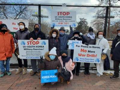 People stand in front of a fence holding signs decrying military exercises in Korea