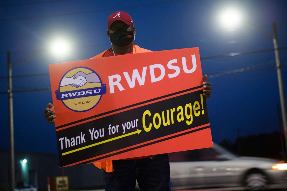 A woker in an orange vest holds a sign reading "RWDSU: THANK YOU FOR YOUR COURAGE" during a roadside demonstration