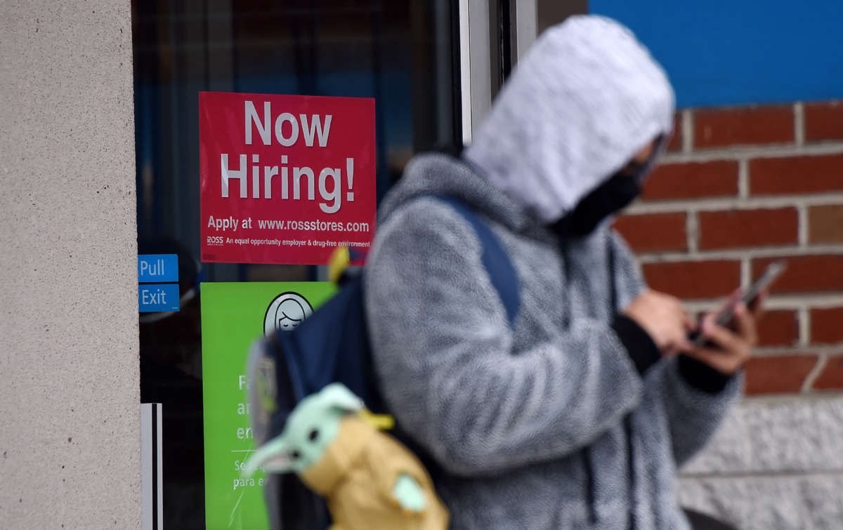 A man wearing a face mask stands next to a "Now Hiring" sign in front of a store on December 18, 2020, in Arlington, Virginia.