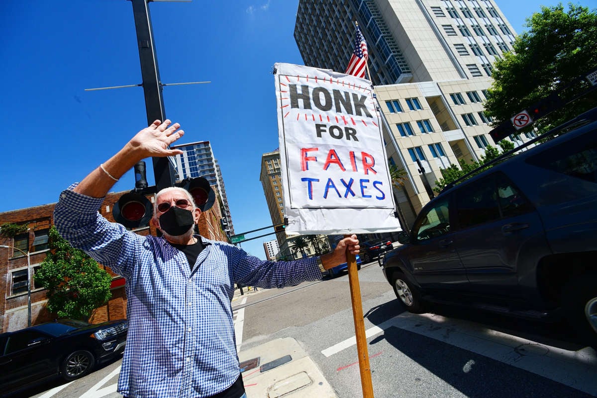 A rad old dude holds a sign reading "HONK FOR FAIR TAXES" during a roadside protest