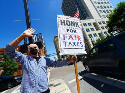 A rad old dude holds a sign reading "HONK FOR FAIR TAXES" during a roadside protest